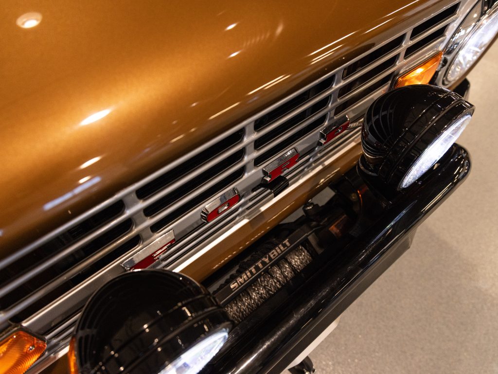 Close-up of a vintage car's front grille and emblem with auxiliary round headlights. - Monarch Showroom