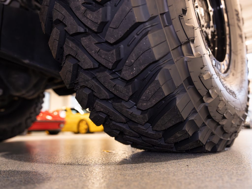 Close-up of rugged off-road vehicle tires with a classic red car in the background. - Monarch Showroom