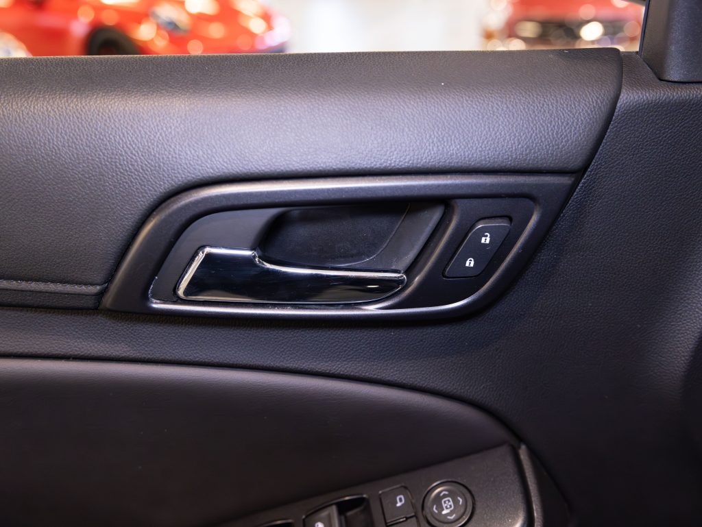 Close-up of a car door's interior handle, featuring a chrome finish and lock/unlock buttons. The black leather panel adds texture, with parts of a red car visible in the background. - Monarch Showroom