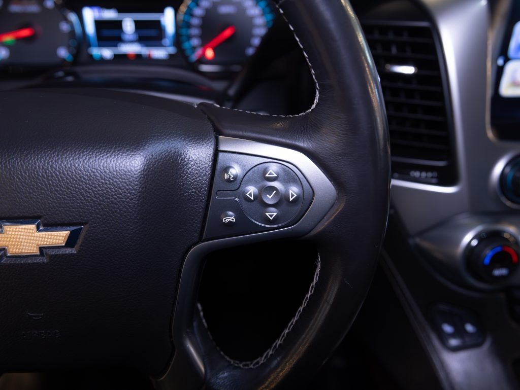 Close-up of a black Chevrolet steering wheel with control buttons. Dashboard with illuminated gauges visible in the background. - Monarch Showroom