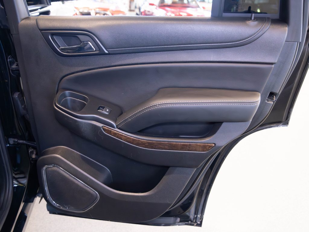 The image shows the interior of a car's rear door, featuring a black leather panel with a wood trim, armrest, speaker, and window control buttons. - Monarch Showroom