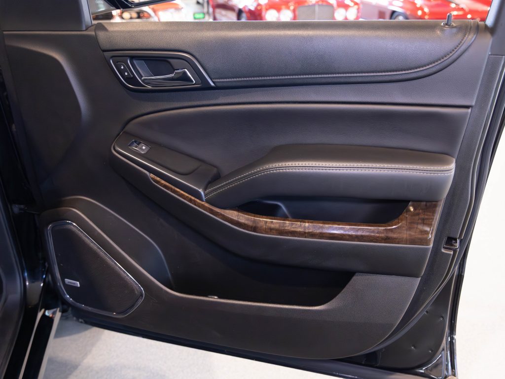 Close-up of a car interior door panel with black leather and wood accents, featuring a door handle, window control buttons, and speaker. - Monarch Showroom