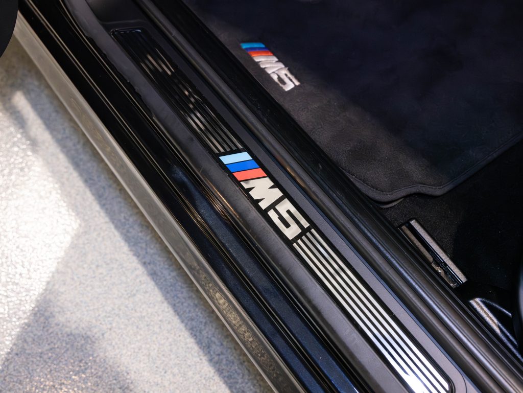 Close-up of a car door sill with the "M" logo in blue, purple, and red on a black background. Another "M" logo is visible on the floor mat inside. - Monarch Showroom